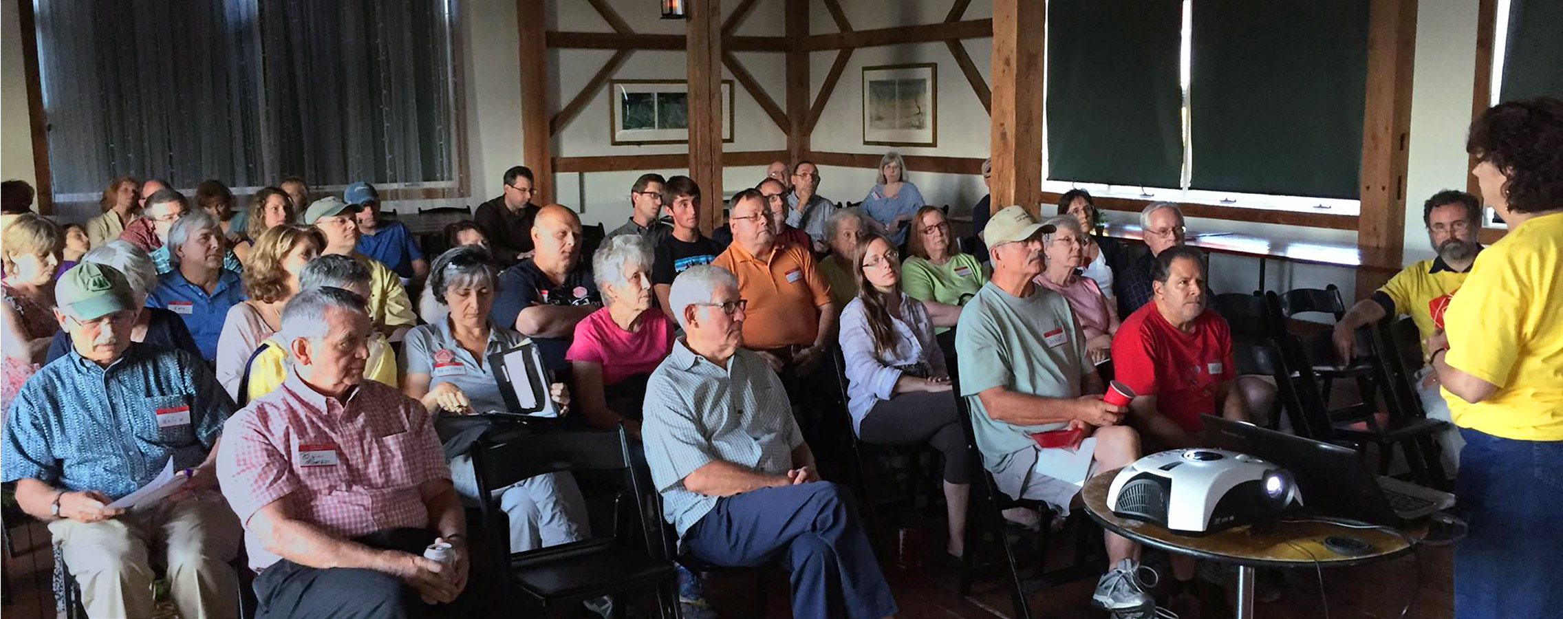A packed room of residents in folding chairs listening to a speaker in a yellow shirt at a community meeting in a timber-framed barn