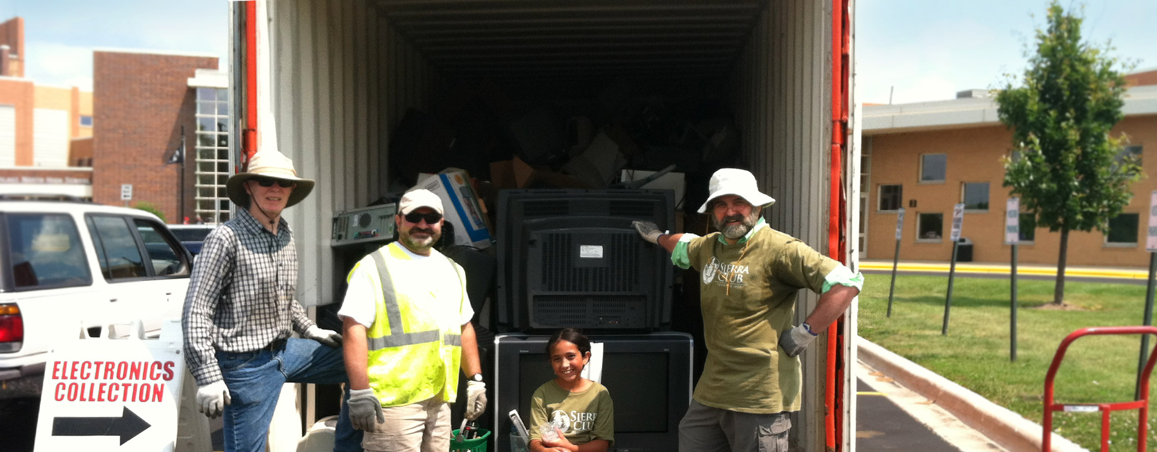 Volunteers in work gloves and sun hats stand at the open back of a trailer full of old televisions and computers at an electronics collection
