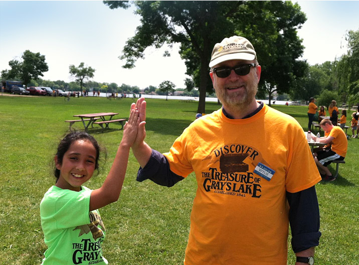 A man in an orange Discover the Treasure of Grayslake T-shirt high-fives a girl in a green shirt at an outdoor event in a park
