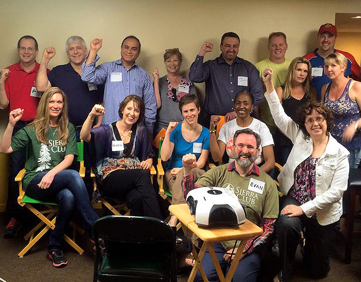 A smiling group of about a dozen volunteers, several raising fists, gathered around a table at a meeting