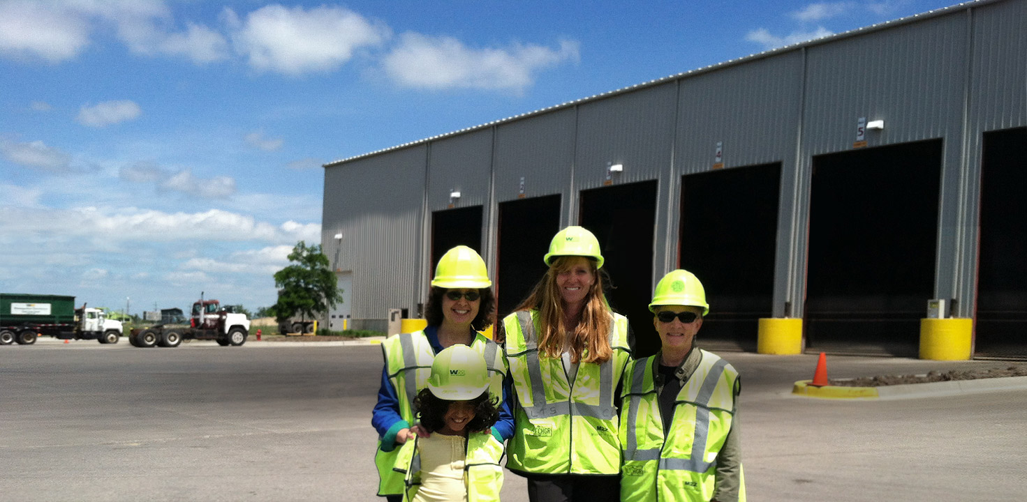 Four visitors in hard hats and safety vests, including a child, smile outside the loading bays of a waste and recycling facility